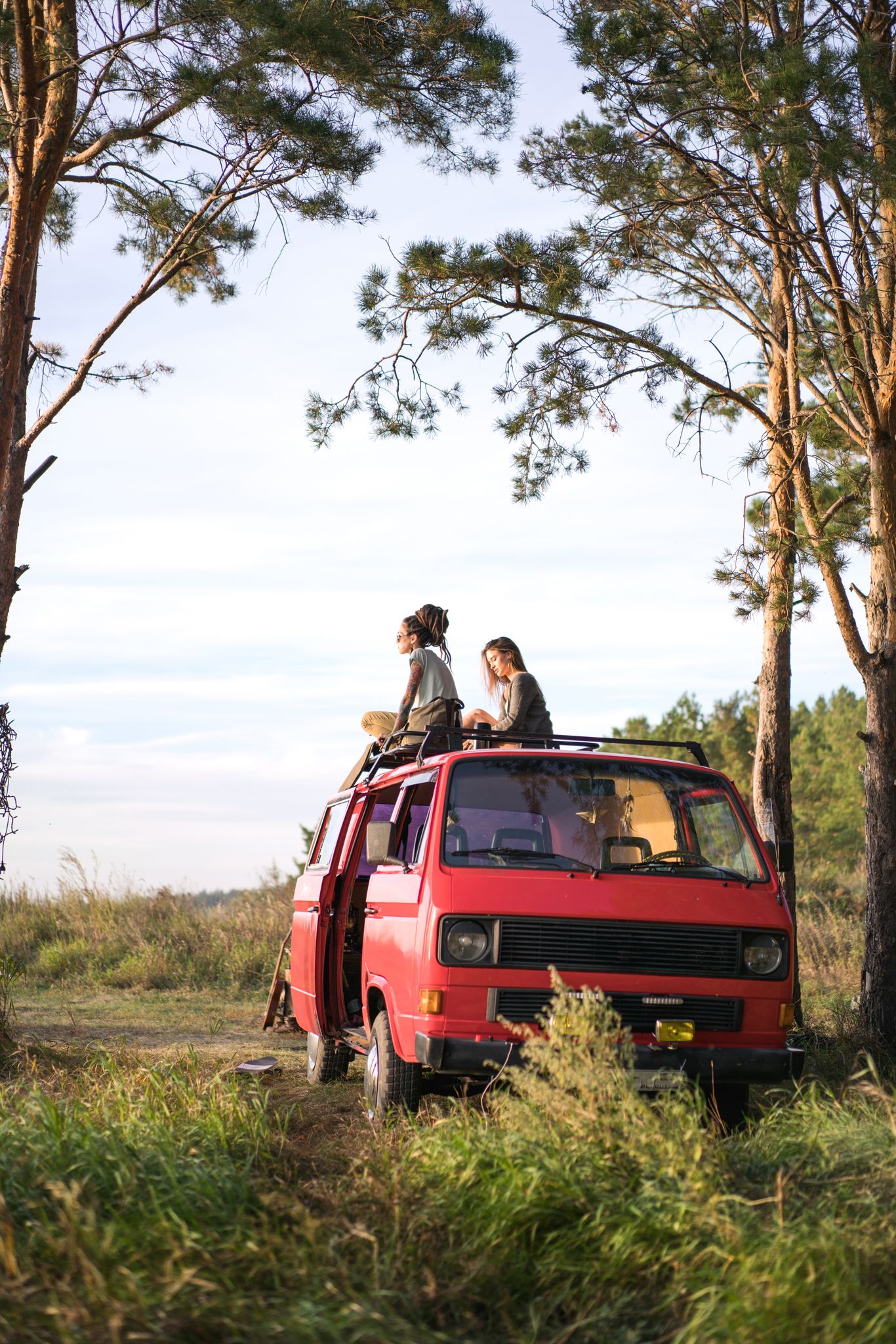 Two women enjoy a scenic view sitting on a red van during a road trip through lush greenery.